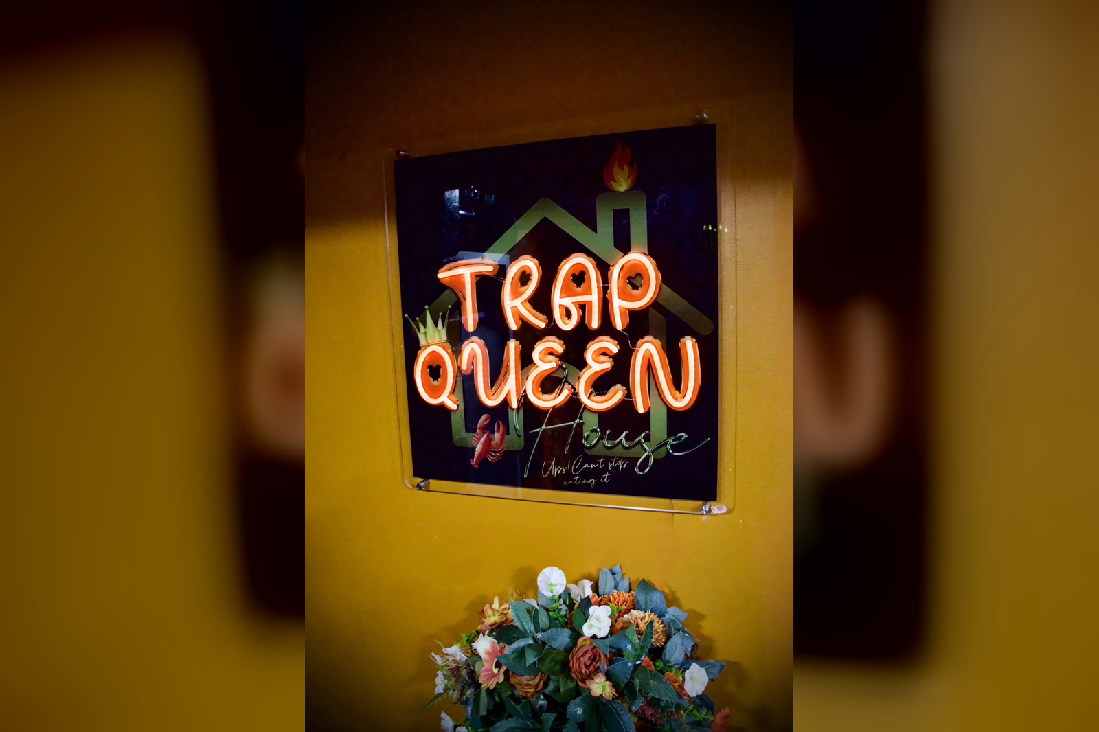 The Trap Queen House neon sign glowing orange on the wall above a bunch of flowers.